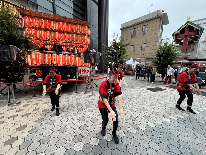 写真：桐生八木節まつりin浅草　桐生八木節キャンペーンスタッフが櫓の前で、両腕を前に揃えて出し、手踊りをしている様子