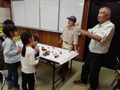 写真:採取したキノコを並べたテーブルの前で子どもたちが講師の話を聞いている